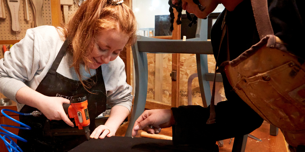 A volunteer at Petit Miracles being taught how to use an upholstery air stapler by the in-house expert, highlighting hands-on skill development and sustainable upcycling practices