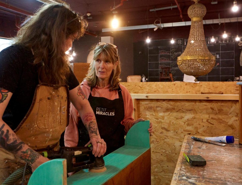 A volunteer at Petit Miracles, a furniture upcycling charity, learning how to use an electric sander under the guidance of the team, showcasing skill-building and sustainability in action.